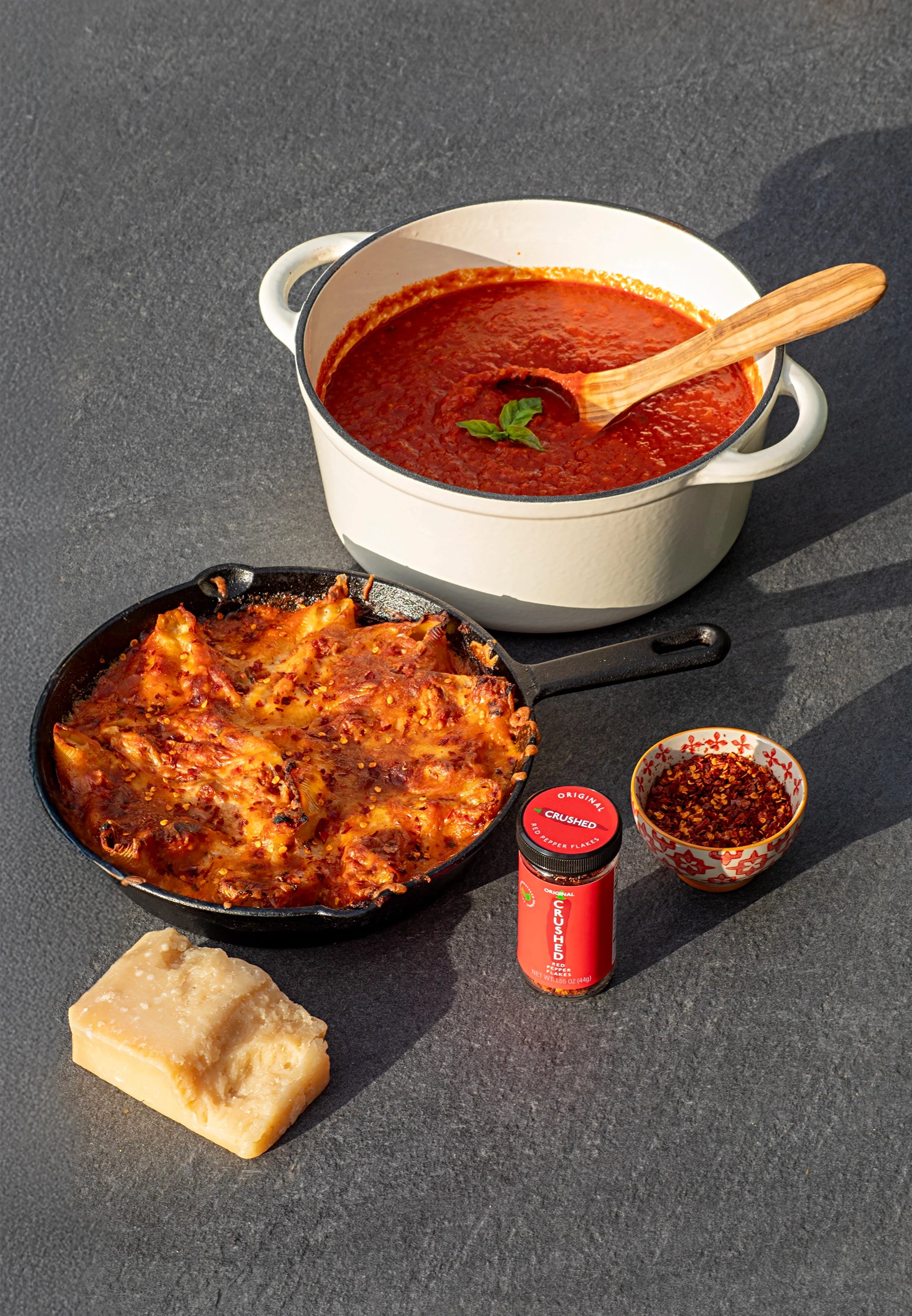 Baked Italian Stuffed Shells in a cast iron skillet alongside a large white dutch oven filled with marinara sauce. A large chunk of Parmesan cheese and Original Crushed Red Pepper Flakes accompany the dish 