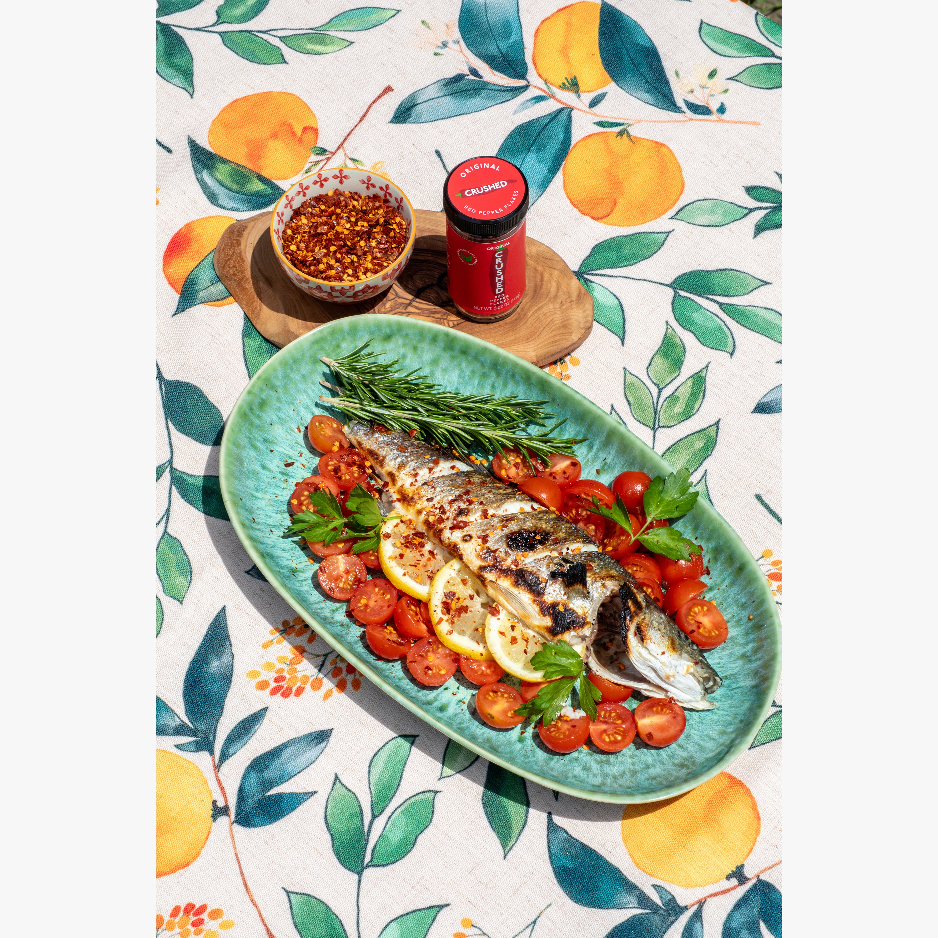 Original Crushed Red Pepper Flakes on a platter of roasted branzino with cherry tomatoes, lemon and rosemary on a decorative Italian table cloth