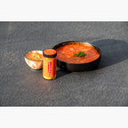 Jar of Cheddar Crushed Red Pepper with a bowl of tomato soup and a bowl of Cheddar Crushed Red Pepper on a slate surface