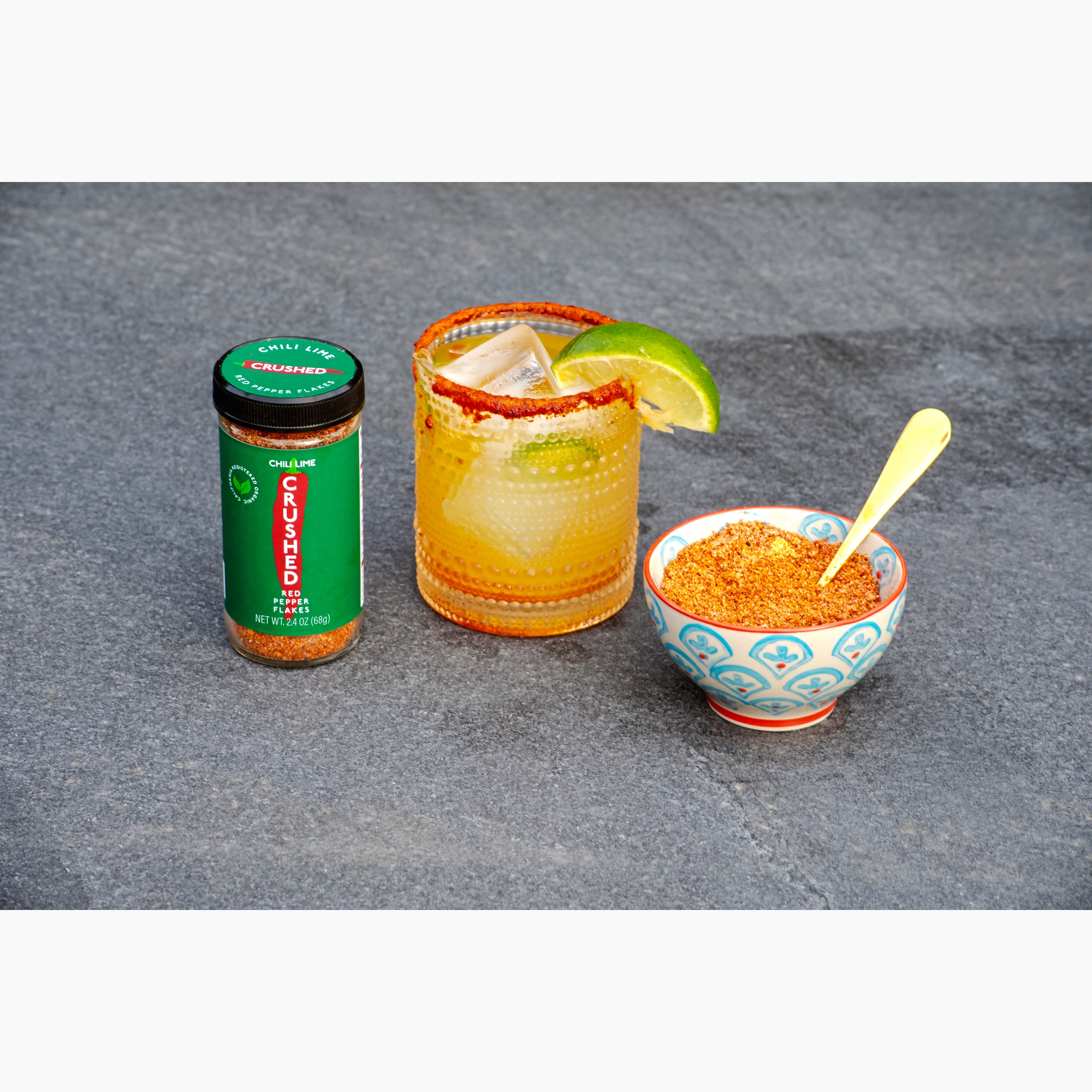 Jar of Chili Lime Crushed Red Pepper,  margarita cocktail with lime and chili lime spice rim, and bowl of Chili Lime Crushed Red Pepper on a gray slate surface.