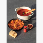 Dishes with red tomato sauce, a skillet with stuffed shells, a chunk of parmesean,  and a jar of Original Crushed Red Pepper on a gray slate surface