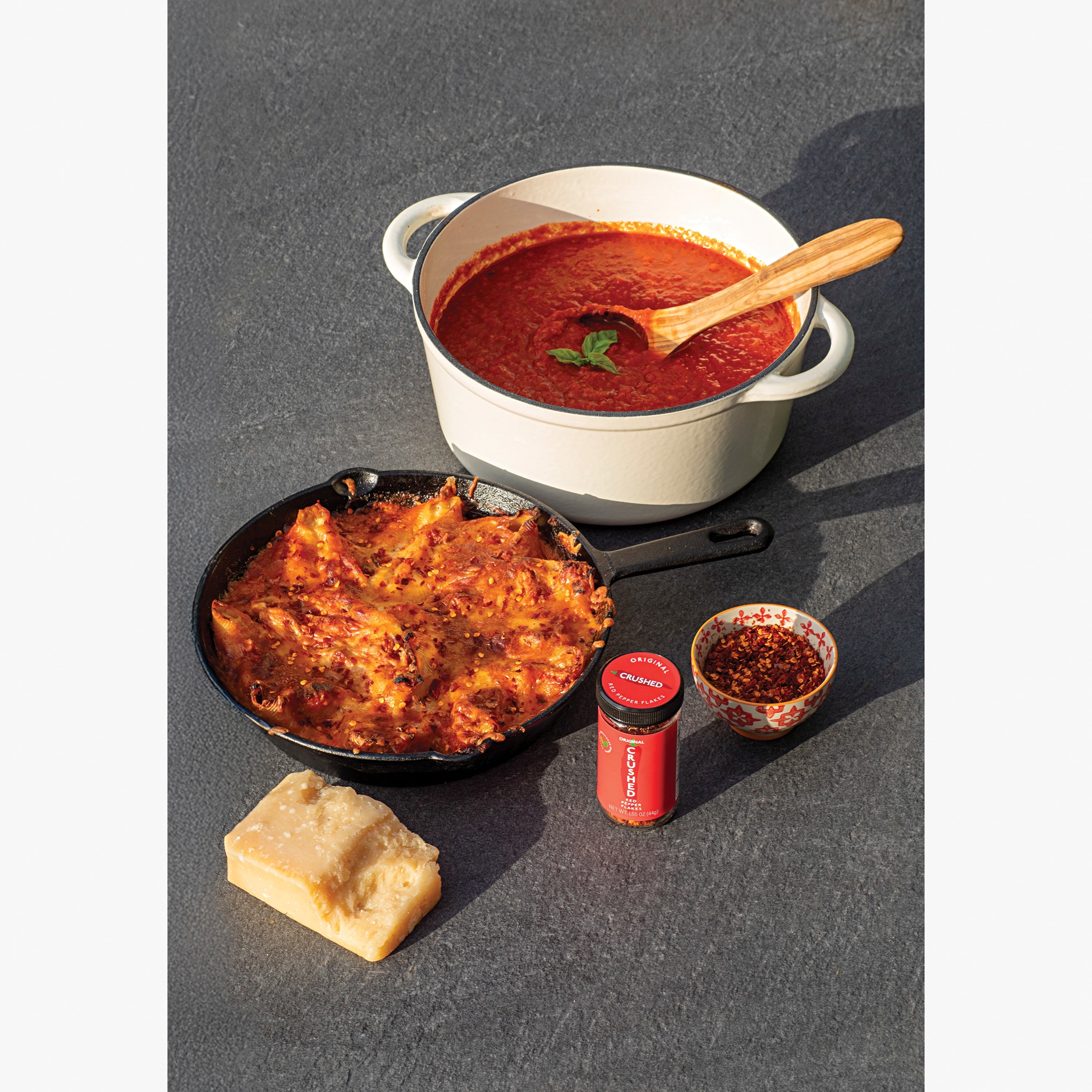 Dishes with red tomato sauce, a skillet with stuffed shells, a chunk of parmesean,  and a jar of Original Crushed Red Pepper on a gray slate surface